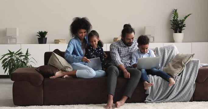 African family and kids sit on sofa using diverse devices in living room at home. Loving parents teach preschool son and daughter spend weekend with modern gadget. Leisure with electronic tech concept
