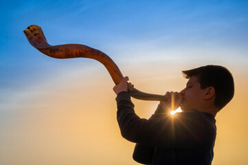 Teen boy blowing Shofar - ram's horn traditionally used for Jewish religious purposes, including the Feast of trumpets, Yom Kippur and Rosh Hashanah; beautiful sunset sky with sunburst in bachground - obrazy, fototapety, plakaty
