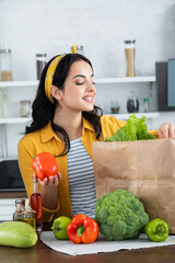 happy brunette woman looking at paper bag with fresh groceries while holding bell pepper