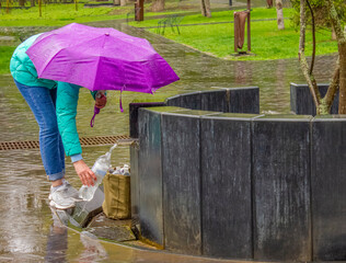 woman with purple umbrella pours water in a plastic bottle from a pump room in a city park on a rainy summer day
