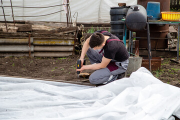 a young guy works at his summer cottage, builds a greenhouse, a screw gun in his hand