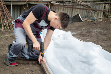 a young guy works at his summer cottage, builds a greenhouse, a screw gun in his hand