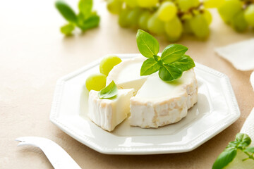 White wheel cheese with basil leaves and grapes. Goat cheese on white plate.