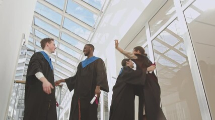 Low angle view of four diverse students wearing university graduate gowns standing in university hall, throwing caps up and then hugging and congratulating each other - Powered by Adobe