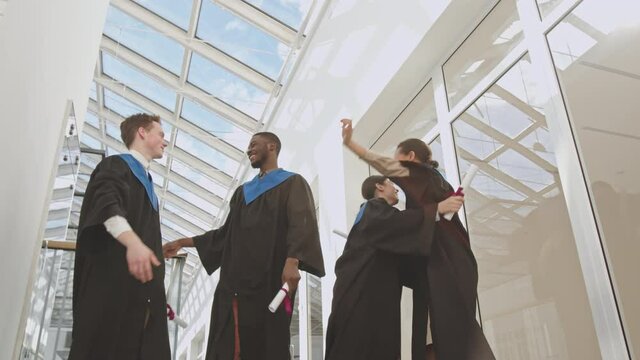 Low angle view of four diverse students wearing university graduate gowns standing in university hall, throwing caps up and then hugging and congratulating each other