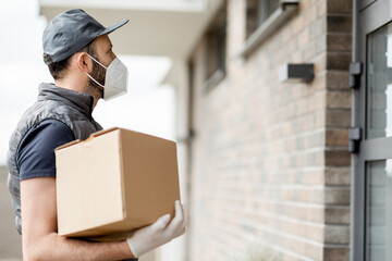 Courier in cap wearing mask and gloves holds a parcel box and staying near front door of house....