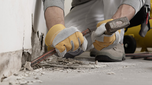 Construction worker hands with gloves working with hammer and chisel to remove old plaster from wall for house renovation, close up with rubble