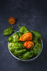 habanero chili peppers, ripe and unripe hot variety of capsicum chinense, green, orange and red color fruits on a bowl isolated on dark texture background
