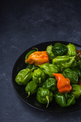 habanero chili peppers, ripe and unripe hot variety of capsicum chinense, green, orange and red color fruits on a plate isolated on dark texture background