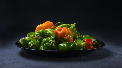 habanero chili peppers, ripe and unripe hot variety of capsicum chinense, green, orange and red color fruits on a plate isolated on dark texture background