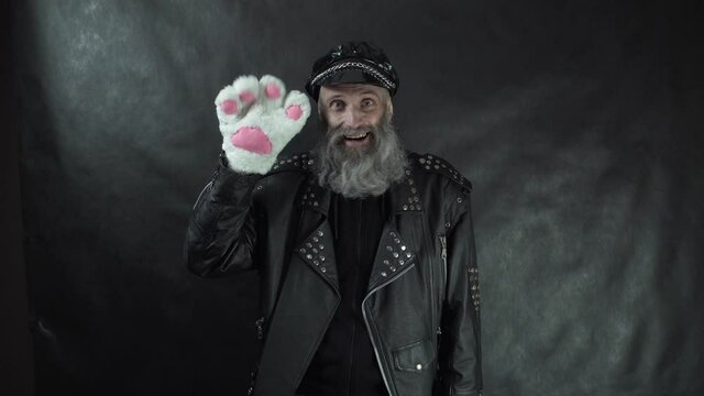 Bearded man in leather jacket and cap smiling sweetly, saying goodbye and waving cat paw glove with black background