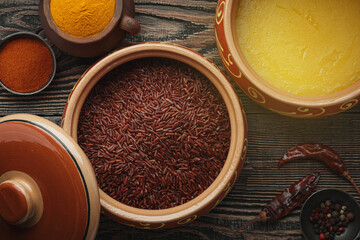 Ghee or clarified butter in ceramic bowls and red rice with different spices on an old wooden table.