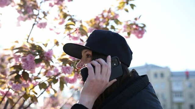 young woman picks up the phone in front of a blossoming sakura in the sun slow motion