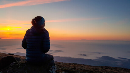 A woman with a headband sitting on top of Babia Gora, Poland, and enjoying the panoramic view on sun rising above the horizon. There are thick clouds below. The sky is pink and orange. Calmness