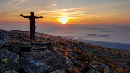 A man in a hoodie standing on top of Babia Gora, Poland, spreading his arms and enjoying the panoramic view on sun rising above the horizon. Thick clouds below. The sky is pink and orange. Freedom