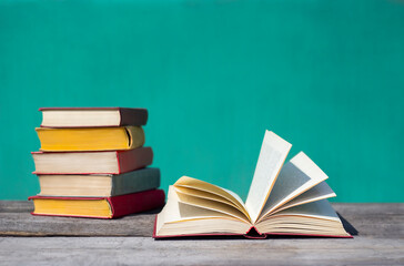 one open book and a stack of colorful books in the background lie against the backdrop of a turquoise school chalkboard in sunlight