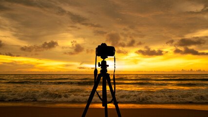 Silhouette of Camera is on a tripod that rests on the sandy beach With a golden background of the sun that falls on the horizon Give you a warm feeling Travel vacation background concept