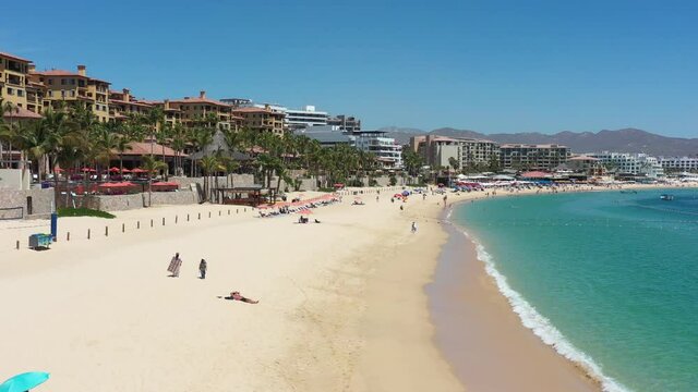 Aerial view over El Medano beach in Cabo San Lucas, Mexico. Drone flying forward with tourists on the beach, a blue sky and the waves crushing over the sand.