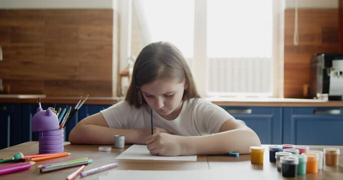 Little kid girl draw a picture with paint on a paper sitting by the table alone in the kitchen. Lifestyle, hobbies, leisure, arts and education concept.