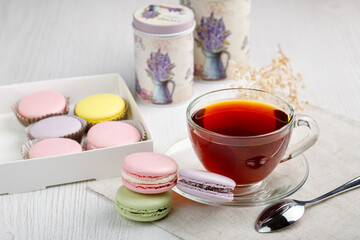 Multicolored macaroons in a box and a cup of tea on a light wood kitchen table.