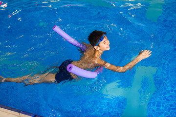 The boy in the pool learns to swim using a stick of foam. The child is fun and cheerful. Top view.