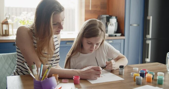 Mother and daughter draw a picture with paint on paper sitting together by the table in the kitchen. Young mom helps her kid to paint a picture. Lifestyle, hobbies, leisure, arts and education concept