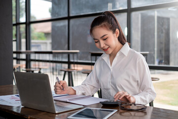Asian businesswoman calculating accounting numbers with a laptop and taking notes at the office.