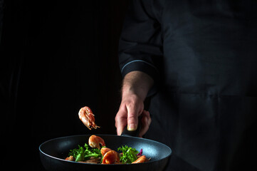 Professional chef or cook cooks shrimp in a pan with vegetables. Cooking seafood, healthy vegetarian food and food on a dark background. Freezing in motion. Free advertising space
