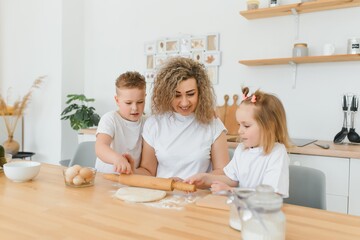 Happy young mother with cute little preschooler kids have fun making dough baking pie or pastry in modern kitchen together, overjoyed mother teach small children doing bakery cooking at home