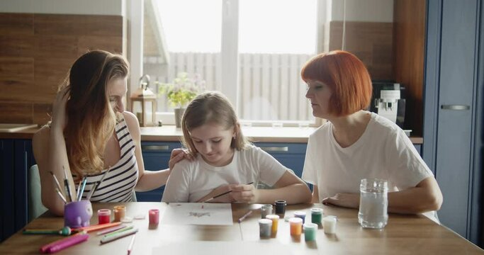 Mother, daughter and grandmother draw a picture with paint on paper sitting together by the table in the kitchen. Lifestyle, hobbies, leisure, arts and education concept
