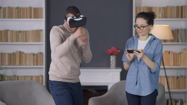Young African American man in VR headset having fun boxing in augmented reality and laughing Caucasian woman using smartphone. Happy interracial couple resting at home on weekend. Lifestyle and unity