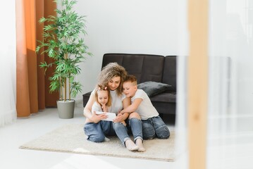 Beautiful young mother, their cute little daughter and son are using a tablet and smiling, sitting on the floor at home
