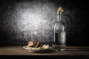 A bottle with a strong drink, and a white plate with a snack, on a wooden table top, on a background with a stain