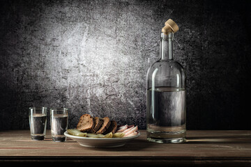 A bottle and two glasses, with a strong drink, and a white plate with a snack, on a wooden table top, on a background with a stain