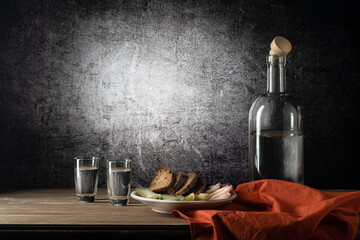 A bottle and two glasses, with a strong drink, and a white plate with a snack, a red napkin, on a wooden table top, on a background with a stain