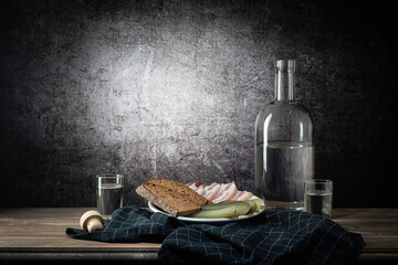 A bottle and two glasses, with a strong drink, and a white plate with a snack, a dark napkin, on a background with a stain