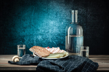 A bottle and two glasses, with a strong drink, and a white plate with a snack, a dark napkin, on a background with a stain