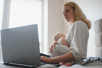 white young mother sitting with baby on bed and working at laptop, work at home