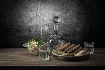 A decanter and two glasses, with a strong drink, with a white plate of snacks (pickles, lard, bread), and an olivier salad, on a wooden table top