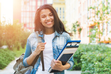Portrait of beautiful happy smiling african american girl student posing outdoors with a backpack and pile of books and tablet computer in hands
