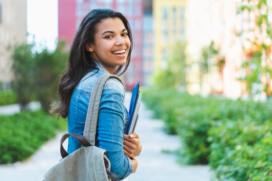 Outdoor portrait of beautiful smiling casually dressed student girl walking with a backpack and pile of books and tablet in hands