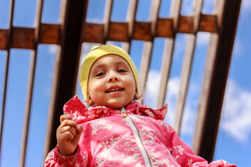 A girl, child in pink overcoat smiling happily against wooden roof and blue sky background. Play outdoors, careless childhood concept