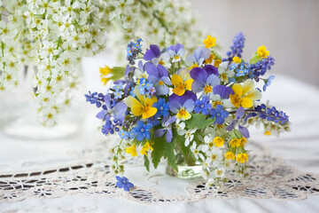 A bouquet of spring blue, yellow flowers in a vase on the table. Pansies, forget-me-nots, primroses, bird cherry, violets, muscari. Postcard, blur, selective focus. - 433510602