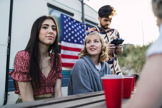 Amigos tomando algo en una mesa de madera frente a una caravana con la bandera americana por el 4 de julio mientras un chico vuela un drone