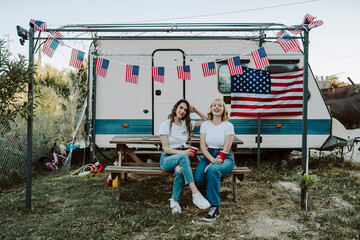 Dos amigas jovenes sentadas frente una caravana con la bandera de estados unidos mientras beben y toman fotos por el dia de la independencia de estados unidos por el 4 de julio - obrazy, fototapety, plakaty