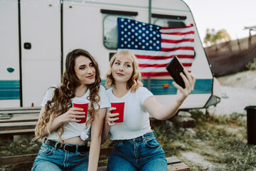 Dos amigas jovenes sentadas frente una caravana con la bandera de estados unidos mientras beben y...