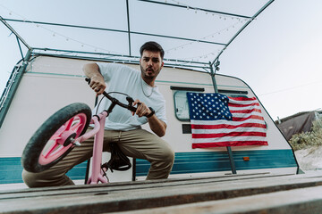 Chico joven con barba sobre mini bici con una caravana de fondo y una bandera de estados unidos