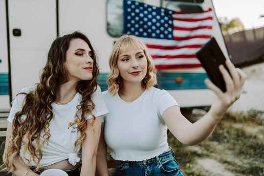 Dos amigas jovenes sentadas frente una caravana con la bandera de estados unidos mientras beben y toman fotos por el dia de la independencia de estados unidos por el 4 de julio