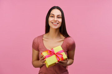 Fototapeta premium Portrait of happy attractive young brunette woman holding gift box, isolated over pastel pink background