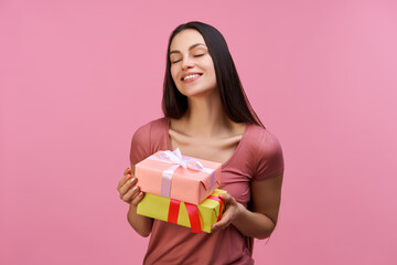 Young brunette woman holding gift boxes with charming smile on her face,  isolated over pastel pink background